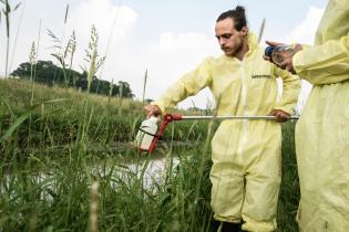 Mann im gelben Anzug nimmt mit einer an einem Stab befestigen Flasche Wasserproben aus einem Fluss.