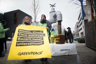 Ein überdimensionierter Wasserhahn, aus dem symbolisch mit Gülle verdrecktes Wasser fließt. Zwei Aktive halten ein Banner: ,Wasser, Klima, Tiere schützen. Bauern dabei unterstützen!