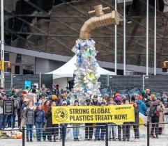 Installation: Aus einem überdimensionierten Wasserhahn tropft Plastik, davor Menschen mit einem Banner "Strong global plastic treaty now".