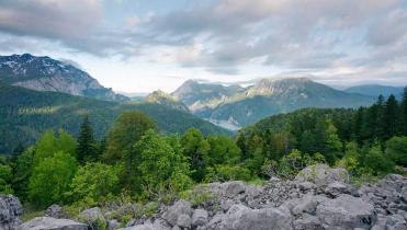 Panorama-Aufnahme des Sutjeska-Nationalparks. Vorn ein Steinhaufen, dahinter ein Meer von Laubbäumen, dann Berggipfel, darüber leicht bewölkter Himmel.