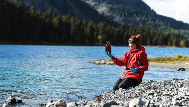 Eine Frau in roter Jacke sitzt an einem See, im Hintergrund sind Bäume zu sehen. Sie hält eine braune Flasche hoch und besieht sich deren Inhalt.