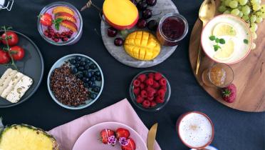 Frühstück mit vielen verschiedenen Obstsorten, Milchkaffee, Käse, Müsli. Appetitlich in Schälchen angerichtet mit Serviette und goldenem Besteck