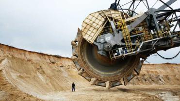 In der Sandgrube eines Tagebaus sieht man das Schaufelrad eines gigantischen Baggers. Daneben steht  winzig ein Mensch mit gelbem Sicherheitshelm.