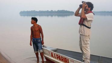 Ein Munduruku steht im glatten Wasser am Ufer des Tapajós, hinter ihm eine baumbestandene Insel, neben ihm ein Ruderbood. Darin steht der Wissenschaftler Luciano Naka; er fotografiert.