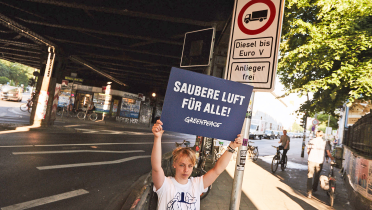 Aktivistin hält Schild „Saubere Luft für alle“ an einer Straßenkreuzung in Hamburg hoch. Im Hintergrund ein Fahrverbotsschild für Diesel-LKW bis Euro-Norm 5.