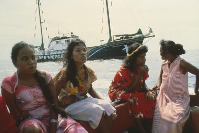 Frauen sitzen in einem Boot, im Hintergrund die Rainbow Warrior