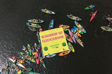 Kajaks, Kanus und Schwimmer:innen im Wasser, auf einem schwimmenden Banner steht: Klimakrise ist Fluchtgrund!