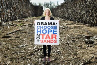 Eine junge Frau steht zwischen zwei riesigen Stapeln aufgeschichteter Baumstämme; sie hält ein Banner mit der Aufschrift "Obama: choose hope not tar sands".