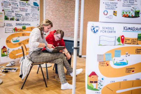 Zwei Menschen sitzen im Klassenraum auf Stühlen und schauen auf einen Laptop