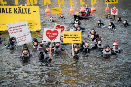 Aktive in Booten und im Wasser der Spree, große Buchstaben auf Schwimmbojen bilden den Schriftzug „Klimageld jetzt!“