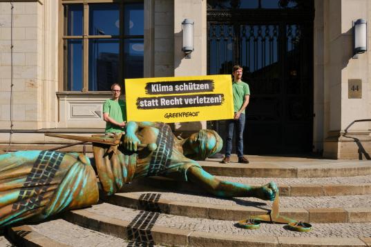 Eine vier Meter große, von Autospuren überzogene Justitia-Figur liegt auf den Stufen zum Verkehrsministerium. Zwei Aktive halten ein Banner: „Klima schützen statt Recht verletzen“.