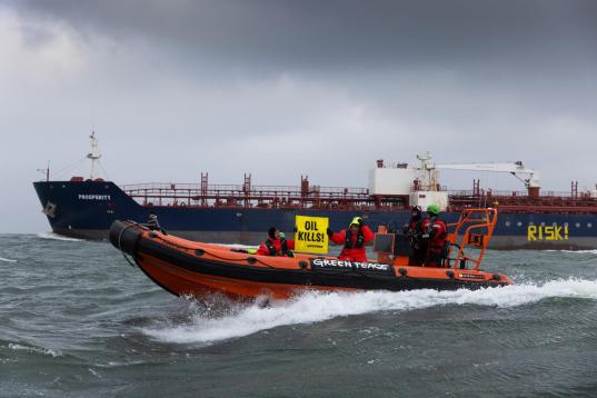 Auf der Tankerwand steht "Risk", davor ein Schlauchboot mit Aktiven, die ein Banner halten "Oil kills".