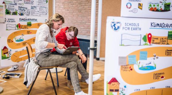 Zwei Menschen sitzen im Klassenraum auf Stühlen und schauen auf einen Laptop