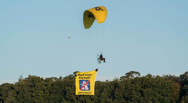 Ein Greenpeace-Aktivist fliegt mit einem elektrisch betriebenen Gleitschirm über den Dannenröder Wald. Am Segler hängt ein Banner mit einem durchgestrichenen Autobahnzeichen und der Forderung „Wald statt Asphalt“.