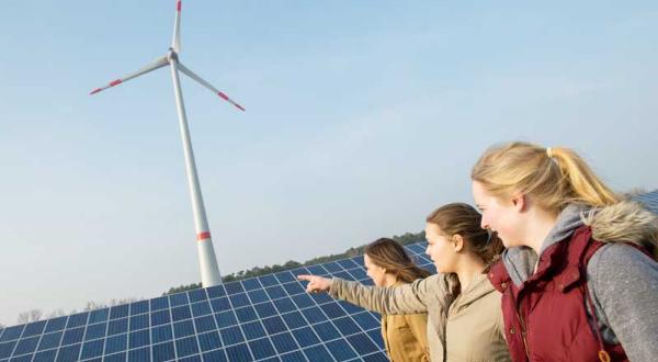 Drei junge Frauen stehen vor einem Solarpanel, dahinter dreht sich ein Windrad. Eine der Frauen zeigt auf die Anlagen.