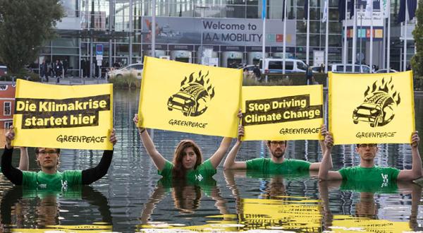 In einem 100 mal 90 Meter großen Wasserbecken stehend, halten Aktivist:innen Banner mit dem Slogan „Die Klimakrise startet hier“ sowie „Stop Driving Climate Change“.