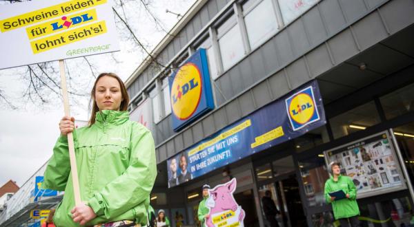 Greenpeace Ehrenamtliche protestieren vor Lidl in Hamburg gegen Billigfleisch. Eine Aktivistin trägt ein Schild mit der Aufschrift „Schweine leiden - für Lidl lohnt sich's!“. Im Hintergrund ein Schwein aus Pappe.