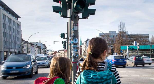 Zwei Schulkinder stehen an einer Straßenkreuzung mit viel Verkehr.