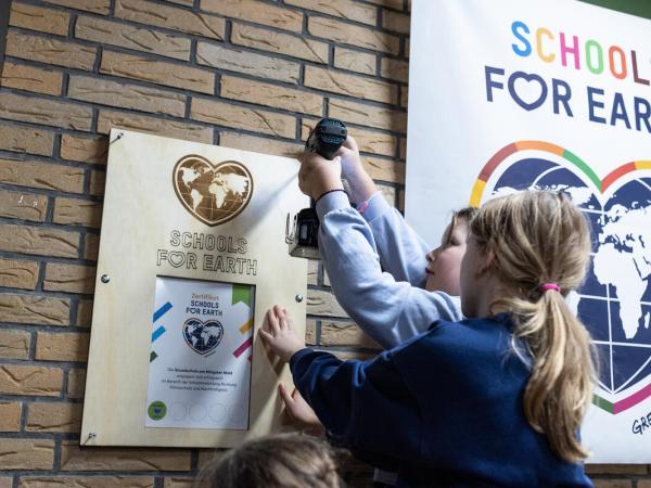 Zwei Mädchen bringen eine Holzplakette an die Wand. Daneben hängt ein Plakat von Schools for Earth