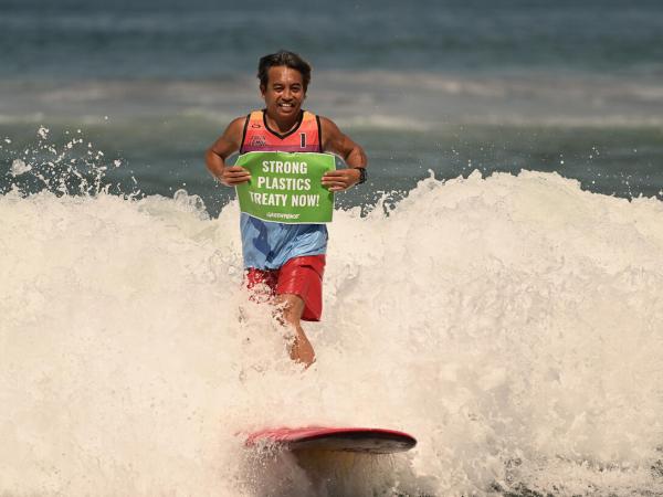 Surfer auf dem Meer hält ein Banner "Strong Plastics Treaty Now!"